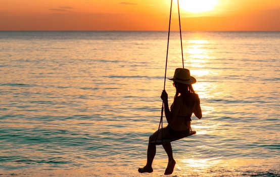 Silhouette woman wear bikini and straw hat swing the swings at the beach on summer vacation at sunset. Enjoying and relaxing girl on holiday. Summer vibes. Woman watching beautiful sunset sky. 