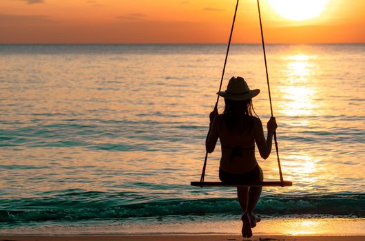 Silhouette woman wear bikini and straw hat swing the swings at the beach on summer vacation at sunset. Girl in swimwear sit on swings and watch beautiful sunset. Summer vibes. Woman travel alone. 