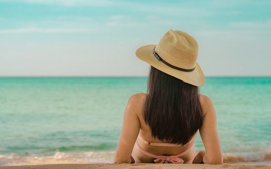 Back view of happy young Asian woman in pink swimsuit and straw hat relax and enjoy holiday at tropical paradise beach at sunset. Girl in summer vacation fashion. Beauty sexy model. Summer vibes.