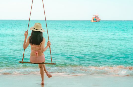 Asian woman wear swimwear and hat swing the swings at sand beach  and looking beautiful tropical paradise sea and sky on sunny day. Summer vacation. Summer vibes. Enjoying and relaxing girl on holiday