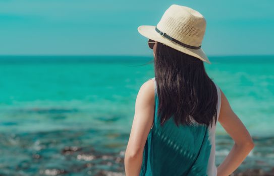 Happy young woman in casual style fashion and straw hat standing at sea beach. Relaxing and enjoy holiday at tropical paradise beach with emerald green water. Girl in summer vacation. Summer vibes.