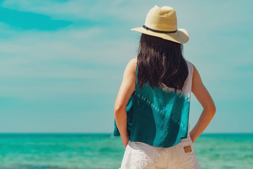 Happy young woman in casual style fashion and straw hat standing at sea beach. Relaxing and enjoy holiday at tropical paradise beach with emerald green water. Girl in summer vacation. Summer vibes.