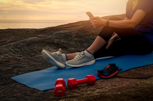 Woman sit and relax on yoga mat near red dumbbells and listen to music via smartphone at stone beach by the sea. Fit girl wear smart band. Healthy, chill out lifestyle. Relax on summer vacations.