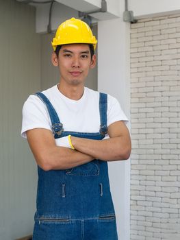 An Asian carpenter wearing a yellow hardhat, standing crossed his arms in the workshop room. 
