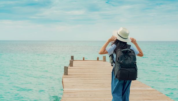 Happy young Asian woman in casual style fashion with straw hat and backpack. Relax and enjoy holiday at tropical paradise beach. Girl stand at the wood pier of resort in summer vacation. Summer vibes.