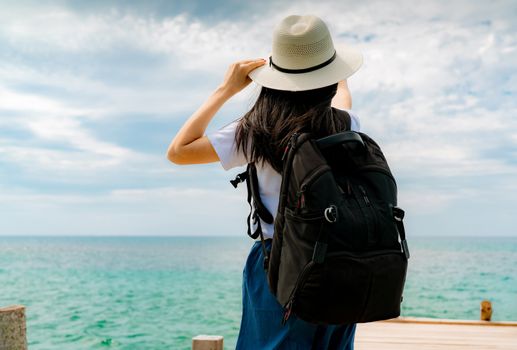 Happy young Asian woman in casual style fashion with straw hat and backpack. Relax and enjoy holiday at tropical paradise beach. Girl stand at the wood pier of resort in summer vacation. Summer vibes.