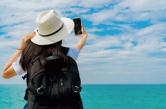 Young Asian backpacker woman wear hat use smartphone taking selfie at pier. Summer vacation at tropical paradise beach. Happy hipster girl travel on holiday. Woman enjoy and relax life. Summer vibes. 