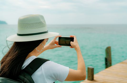 Young Asian backpacker woman wear hat use smartphone taking photo at pier. Summer vacation at tropical paradise beach. Happy hipster girl travel on holiday. Woman enjoy and relax life. Summer vibes. 