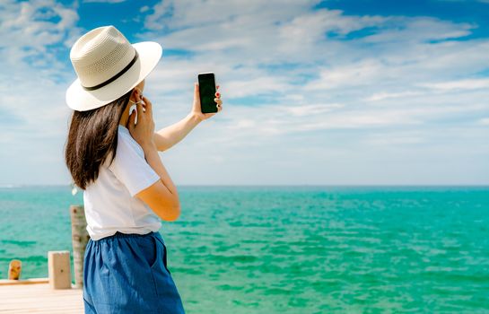 Young Asian woman wear hat in casual style use smartphone taking selfie at pier. Summer vacation at tropical paradise beach. Happy girl travel on holiday. Woman enjoy and relax life. Summer vibes. 