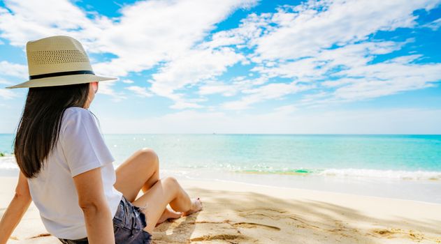 Happy young woman in white shirts and shorts sitting at sand beach. Relaxing and enjoying holiday at tropical paradise beach with blue sky and clouds. Girl in summer vacation. Summer vibes. Happy day.