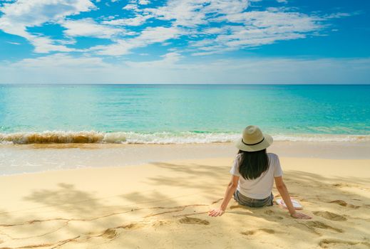 Happy young woman in white shirts and shorts sitting at sand beach. Relaxing and enjoying holiday at tropical paradise beach with blue sky and clouds. Girl in summer vacation. Summer vibes. Happy day.