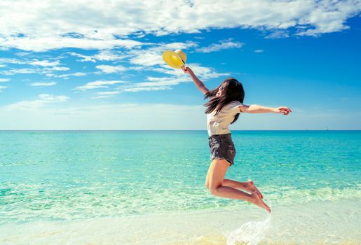 Happy young woman in casual style fashion and straw hat jumping at sand beach. Relaxing, fun, and enjoy holiday at tropical paradise beach with blue sky and white clouds. Girl in summer vacation.