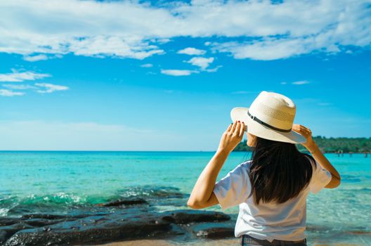 Happy young Asian woman in casual style fashion with straw hat stand at sea beach of resort in summer vacation . Relaxing and enjoying holiday at tropical paradise beach. Summer vibes. Summer travel.