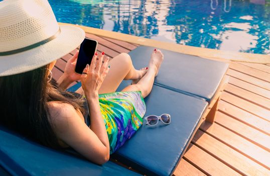 Asian woman with hat and swimsuit sit on sunbed at poolside and using smartphone on summer vacation by swimming pool. Girl with nail manicure and sunglasses. Summer vibes. Woman relaxing on holiday.