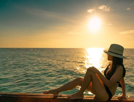Sexy and relax woman wear black bikini with straw hat sit on wooden beam near sand beach at sunset. Girl enjoy holiday at tropical paradise beach on summer vacation. Holiday travel. Summer vibes.