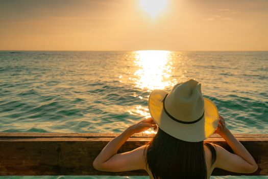 Back view of black long hair sexy Asian woman with straw hat relaxing and enjoying holiday at tropical paradise beach and watch the sunset sky. Summer vacation. Holiday travel alone. Summer vibes. 
