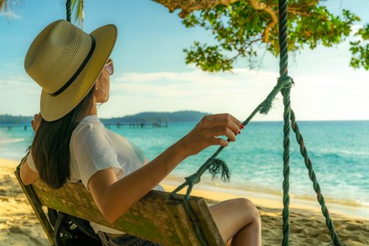 Young Asian woman sit and relax on swings at seaside on summer vacation. Summer vibes. Woman travel alone on holiday. Backpacker wear hat and sunglasses enjoy the day at tropical paradise beach. 