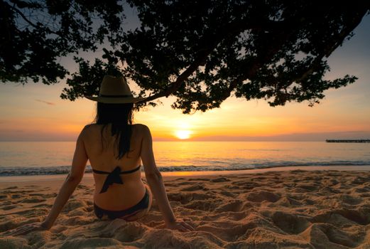 Back view of pregnant woman sit on sand and watching sunset at tropical beach. Woman wear swimsuit and straw hat relaxing at tropical paradise beach on summer vacation. Holiday travel alone.
