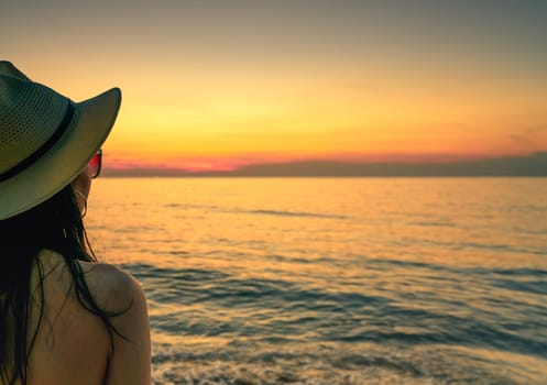 Back view of sexy woman watching sunset at the beach. Woman wear sunglasses and straw hat relaxing at tropical paradise beach. Summer vacation. Holiday travel alone. Summer vibes. Beautiful sunset sky