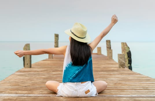 Back view of happy young Asian woman in casual style fashion and straw hat relax and enjoy holiday at tropical paradise beach. Girl stand at the wood pier of resort in summer vacation. Summer vibes.