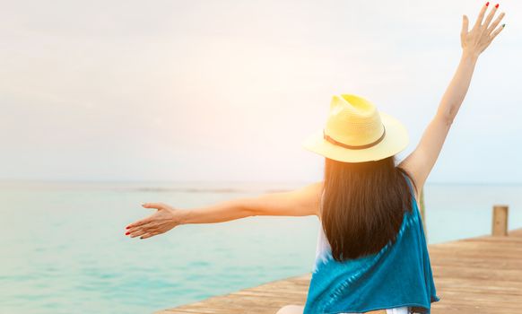 Back view of happy young Asian woman in casual style fashion and straw hat relax and enjoy holiday at tropical paradise beach. Girl stand at the wood pier of resort in summer vacation. Summer vibes.