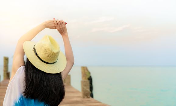 Back view of happy young Asian woman in casual style fashion and straw hat relax and enjoy holiday at tropical paradise beach. Girl sit at the wood pier of resort in summer vacation. Summer vibes.