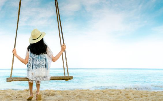 Asian woman in casual style wear hat and sandals sit on swings at sand beach  and looking beautiful tropical paradise sea and sky. Summer vacation. Summer vibes. Enjoying and relaxing girl on holiday.