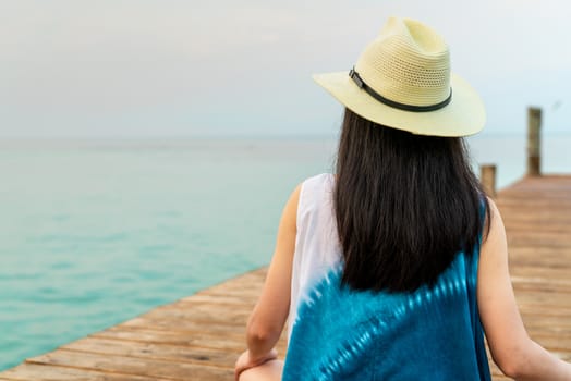 Back view of happy young Asian woman in casual style fashion and straw hat relax and enjoy holiday at tropical paradise beach. Girl stand at the wooden pier of resort in summer vacation. Summer vibes.