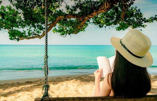 Women sit and reading a book on swings under the tree by the sea. Back view of sexy Asian woman with straw hat relax and enjoy holiday at tropical paradise sand beach. Summer vacation. Summer vibes. 