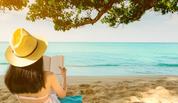Women sit and reading a book under the tree at seaside. Back view of sexy Asian woman with straw hat relaxing and enjoying holiday at tropical paradise sand beach. Summer vacation. Summer vibes. 