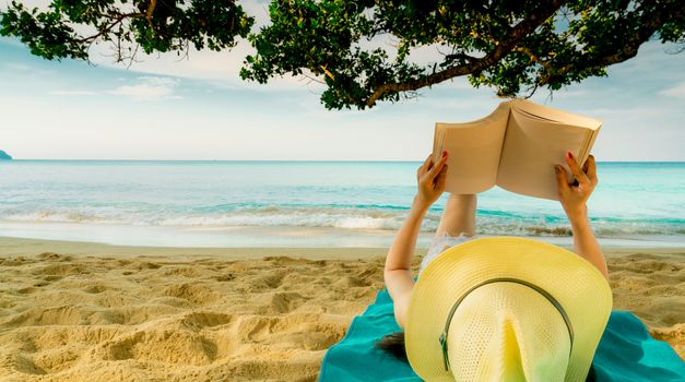 Woman lie down on green towel that put on sand beach under the tree and reading a book. Slow life on summer vacation. Asian woman with hat relaxing and enjoying holiday at tropical beach. Summer vibes