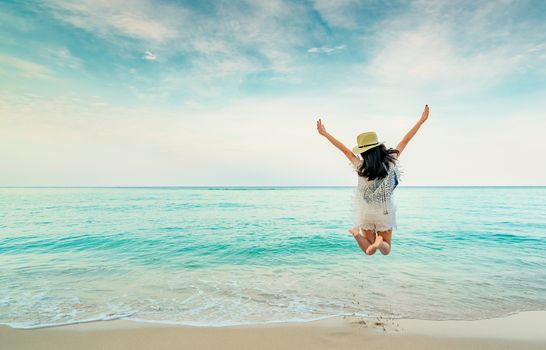 Happy young woman in casual style fashion and straw hat jumping at sand beach. Relaxing, fun, and enjoy holiday at tropical paradise beach with blue sky and white clouds. Girl in summer vacation.