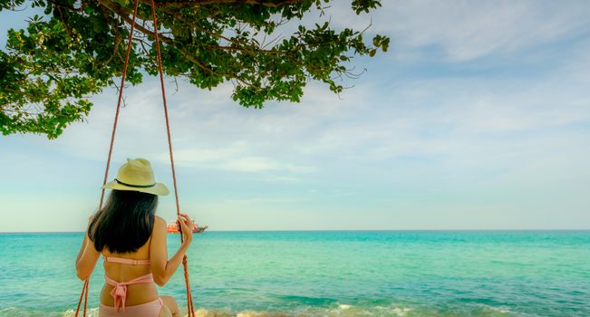 Asian woman wear swimwear and hat swing the swings at sand beach  and looking beautiful tropical paradise sea and sky on sunny day. Summer vacation. Summer vibes. Enjoying and relaxing girl on holiday