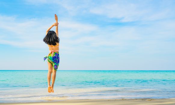 Happy young woman in swimsuit jumping at sand beach. Relaxing and enjoying holiday at tropical paradise beach with blue sky and clouds. Girl in summer vacation. Summer vibes. Happy day. Travel alone.