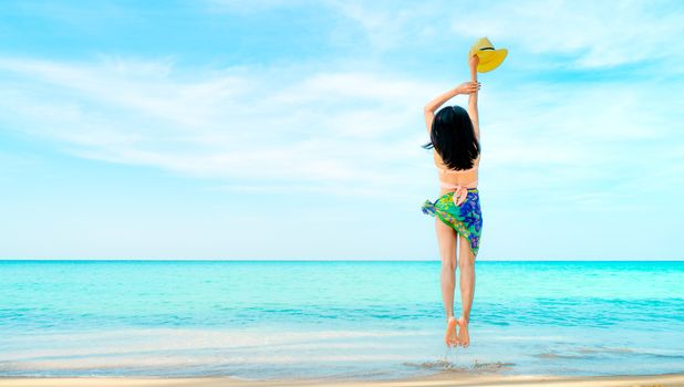 Happy young woman in pink swimwear hand holding hat and jumping at sand beach. Relaxing and enjoying holiday at tropical paradise sea beach. Girl in summer vacation. Summer vibes. Traveling alone.