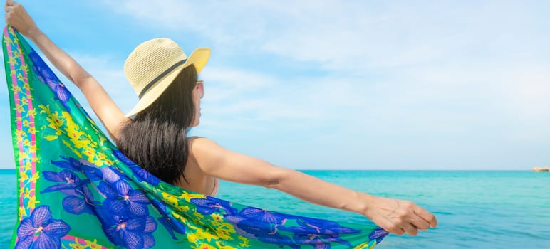 Back view of Asian woman wear swimsuit and opened arms at tropical beach on sunny day with beautiful blue sky and white clouds. Woman travel on summer vacation. Summer vibes. Happy girl.