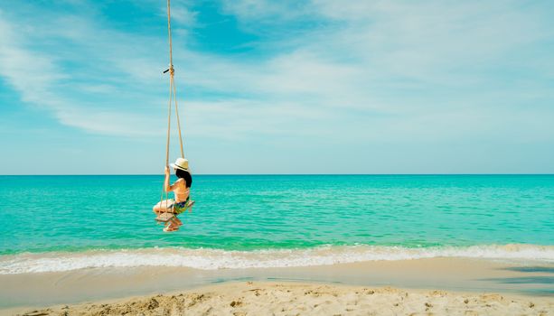 Asian woman wear swimwear and hat swing the swings at sand beach  and looking beautiful tropical paradise sea and sky on sunny day. Summer vacation. Summer vibes. Enjoying and relaxing girl on holiday