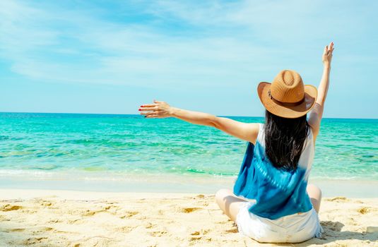 Happy young woman wear straw hat sit and raised hand at sand beach. Relaxing and enjoy holiday at tropical paradise beach with green water. Girl in summer vacation. Summer vibes. Carefree concept.