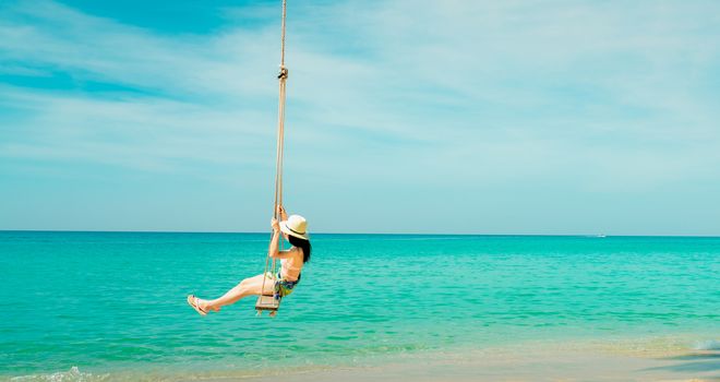 Happy young Asian woman in pink swimsuit and straw hat relax and enjoy holiday at tropical paradise beach. Woman fun on swing. Girl in summer vacation fashion. Green sea water and blue sky and clouds.