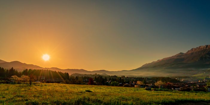 Morning sunrise over the mountain with clear blue sky. Grass field and pine forest in rural village near rock mountain. The fog covered the mountain in the morning. City in valley. Landscape meadow 