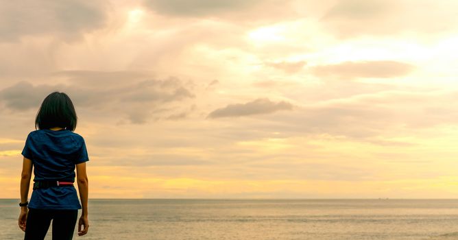 Asian woman watching sunrise sky at sea beach. Runner relax after running at tropical beach. Healthy lifestyle. Girl travel alone on summer vacation. Holiday at the coast of the ocean. Happy woman.