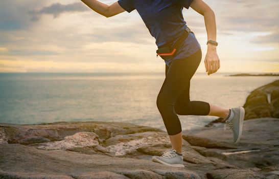 Woman runner running at stone beach by the sea in the morning with sunrise. Fit and strong healthy woman outdoor workout. Fit girl wear smart band and waist bag. Woman running for cardio exercise.