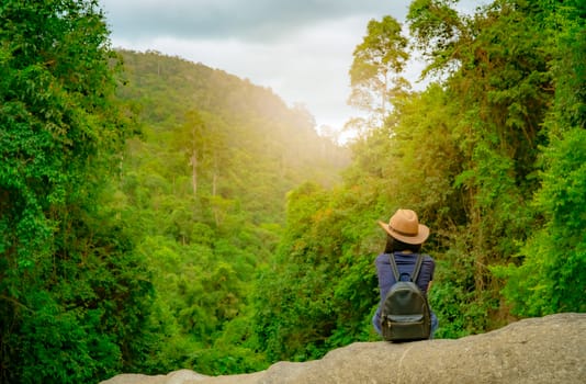 Happy woman travel alone in woods. Active woman with good mood sit on stone in green valley with dense tree in the forest. Happy vacation. Back view of happy woman wear backpack, smart band, and hat.