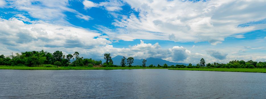 Beautiful landscape view of lake in front of the mountain with blue sky and white cumulus clouds. Green tree and grass field around pond. Tropical weather in summer. Nature landscape. Fresh air.