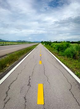 Perspective cracked asphalt road ahead to the mountain. Long straight damaged asphalt road. Bad surface of bike lane and white sky and clouds with green grass beside the empty road. Nature landscape.