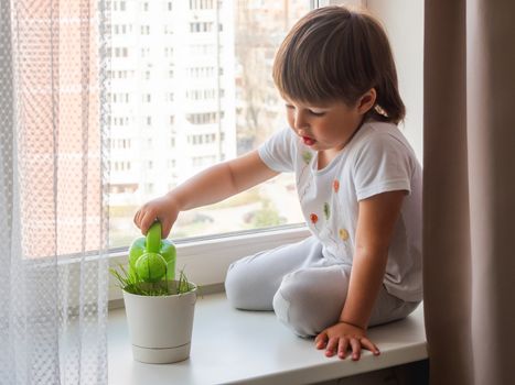 Toddler boy sits on windowsill and waters green grass in flower pot. Little child with green watering can. Kid's first first duties at home.