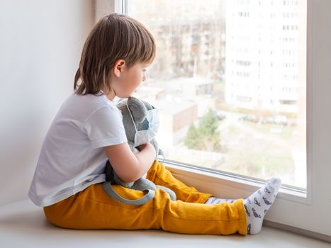 Toddler boy sits with teddy bear in medical mask. Kid with plush toy look through window outside. Koala bear and child on home quarantine because of coronavirus COVID-19.
