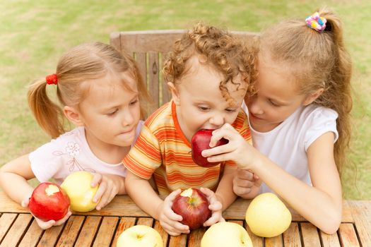 three happy children sitting at the table and eat apples