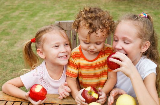 three happy children sitting at the table and eat apples