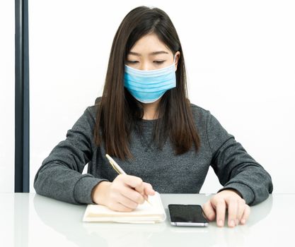 Teenage female student preparing for learnning at home sitting at a desk in front of smartphone with notepad and wear protective medical mask for protect Covid-19 or corona virus disease